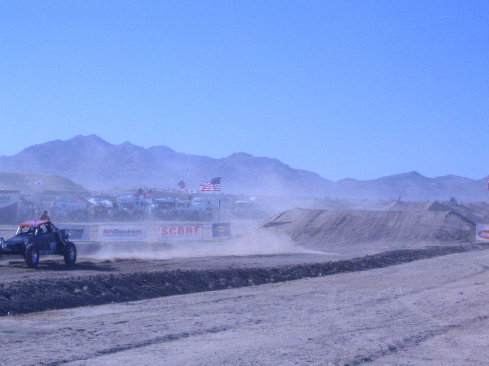 Off-road buggy racing past a dirt berm in a dusty desert with spectators