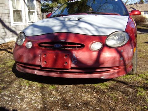 1997 Ford Taurus - The front of a red 1996 Ford Taurus with a white hood and a damaged bumper is parked outside.