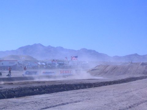 1990 Ford Bronco - dusty desert off-road race course with a dirt berm, American flags, and SCORE and BFGoodrich banners beneath distant mountains.