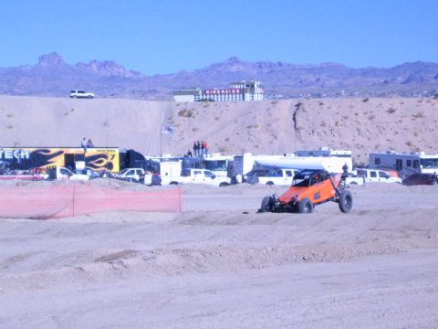1990 Ford Bronco - orange off-road race buggy sits on a sandy desert track in front of a FABTECH trailer, support trucks, and the Riverside resort.