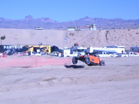 1990 Ford Bronco - orange off-road race buggy climbs a sandy berm behind orange fencing, with a FABTECH trailer and the Riverside resort in the distance.