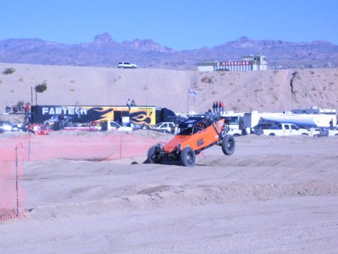 1990 Ford Bronco - orange off-road race buggy drives a sandy desert course past orange fencing, with a FABTECH trailer, support trucks, and a hotel behind.