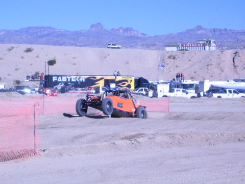 1990 Ford Bronco - orange off-road race buggy rounds a sandy desert track behind orange fencing, with a FABTECH semi-trailer and support trucks behind.