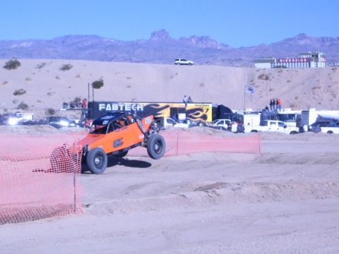 1990 Ford Bronco - orange off-road race buggy sits behind orange course fencing with a FABTECH trailer, support trucks, and spectators in a desert pit area.