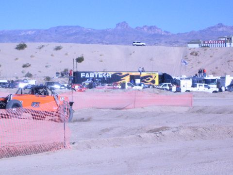 1990 Ford Bronco - front of an orange off-road buggy peeks past orange fencing, with a FABTECH trailer and a busy desert pit area in the background.