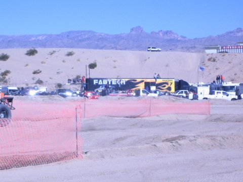1990 Ford Bronco - desert race pit area with a FABTECH semi-trailer, support trucks, and spectators behind orange course fencing under blue sky.