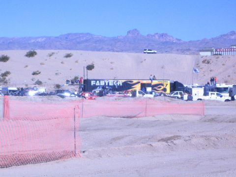 1990 Ford Bronco - FABTECH semi-trailer and a row of support vehicles fill a desert race pit area behind orange course fencing beneath rugged mountains.