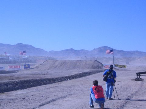 1990 Ford Bronco - Two photographers, one kneeling in a red vest, shoot toward a distant dirt jump on a dusty desert race course lined with flags and banners.