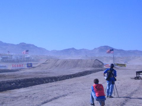 1990 Ford Bronco - Two trackside photographers face a desert dirt jump as American flags and SCORE and Bilstein banners line the dusty off-road course.
