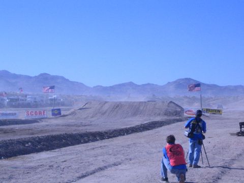 1990 Ford Bronco - Two photographers wait beside their gear watching an empty desert dirt jump, with American flags and sponsor banners along the course.