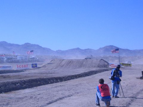 1990 Ford Bronco - Two trackside photographers shoot a dusty desert race jump framed by American flags and SCORE and Bilstein banners under blue sky.