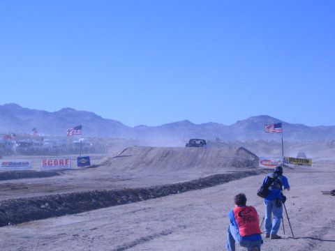 1990 Ford Bronco - Two photographers shoot toward a distant truck cresting a dirt jump on a dusty desert course lined with American flags and sponsor banners.