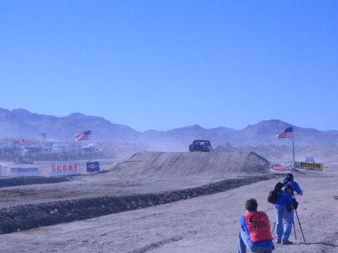 1990 Ford Bronco - distant race truck sits atop a desert dirt jump as two photographers shoot from the dusty foreground past flags and sponsor banners.