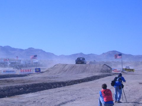 1990 Ford Bronco - race truck crests a desert dirt jump amid heavy dust while two photographers shoot from the foreground beside American flags and banners.
