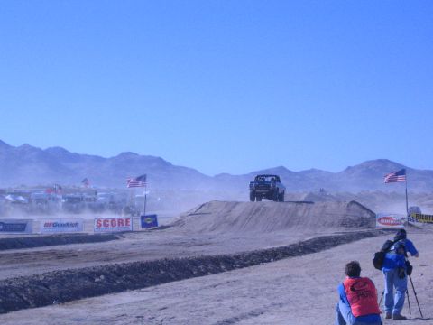 1990 Ford Bronco - race truck crests a desert dirt jump trailing dust while a kneeling photographer in a red vest watches near American flags and banners.