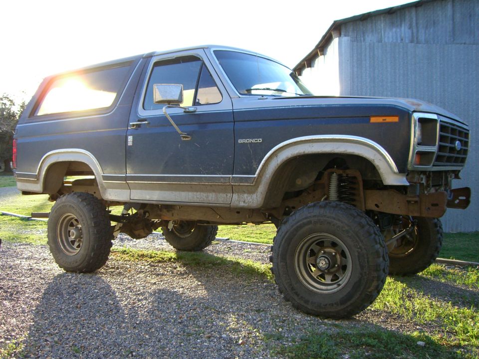 Lifted dark blue Ford Bronco SUV on big off-road tires, side view, parked on gravel by a metal barn