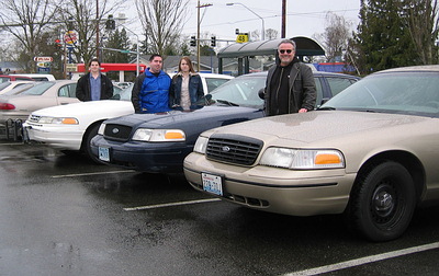1999 Ford Crown Victoria - Several Ford Crown Victoria sedans are lined up in a parking lot with people standing next to them.
