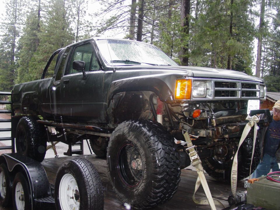 Dark green pickup truck with oversized tires on a trailer, front 3/4 view