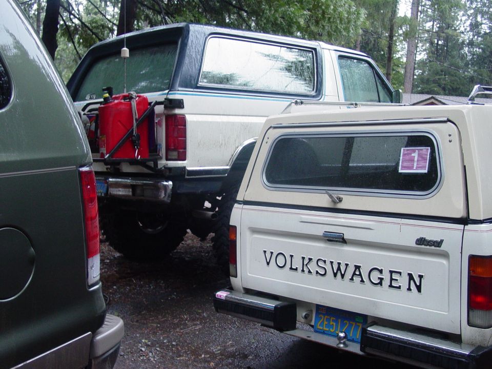 White Ford Bronco with gas can on back, parked next to a Volkswagen pickup truck. Green van on left