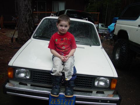 1986 Ford Bronco - A child sits on the hood of a white car, with another vehicle visible to the right.