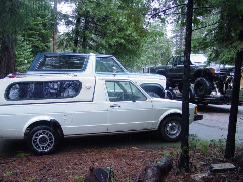 1986 Ford Bronco - A white pickup truck with a camper shell is parked in front of a Ford Bronco and a lifted truck on a trailer.