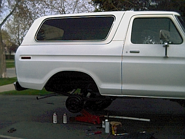 Side view of a white 1978 Ford Bronco with its rear wheel off and a jack underneath