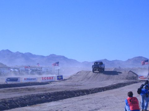 1990 Ford Bronco - off-road race truck launches over a dirt berm on a desert course lined with American flags, SCORE and BFGoodrich banners.