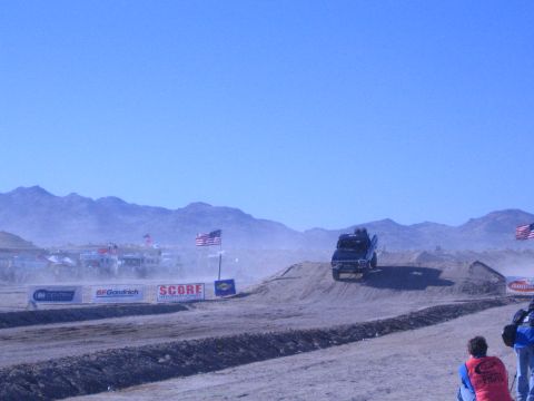 1990 Ford Bronco - dark off-road race truck goes airborne over a dirt jump amid dust, with desert mountains and sponsor banners behind.