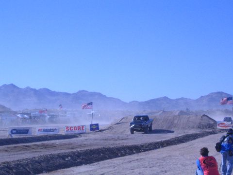 1990 Ford Bronco - blue off-road race truck races along a dusty desert course backed by mountains, flags and sponsor banners.