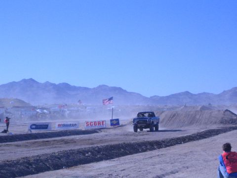 1990 Ford Bronco - off-road race truck runs a desert course past an American flag, sponsor banners and red-shirted course workers.