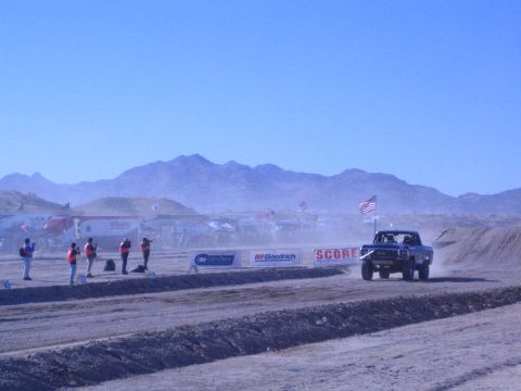 1990 Ford Bronco - blue off-road race truck runs a desert track as course workers in vests line the edge near sponsor banners.