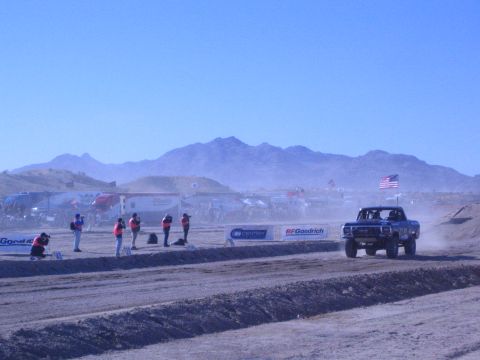 1990 Ford Bronco - blue off-road race truck races a dusty desert course with photographers and workers along the edge and mountains beyond.