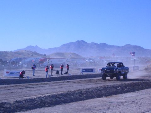 1990 Ford Bronco - blue off-road race truck speeds along a desert track past course workers, a sponsor banner and the pit area.