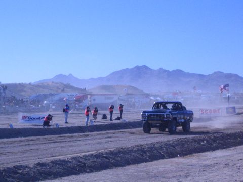 1990 Ford Bronco - blue off-road race truck runs a dusty desert course past workers and a SCORE banner with support trucks behind.