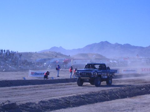 1990 Ford Bronco - blue off-road race truck corners on a dusty desert track past a BFGoodrich banner and spectators.