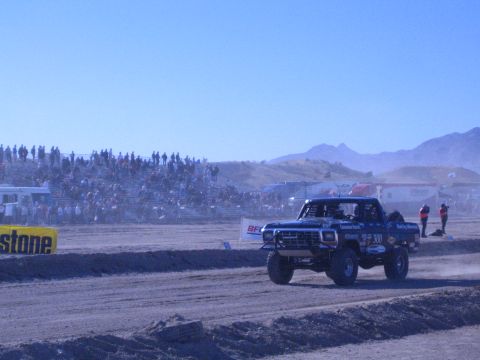 1990 Ford Bronco - Blue off-road race truck number 300 runs the desert track in front of packed grandstands, with a Firestone banner at left.