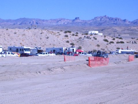 1990 Ford Bronco - RVs and trucks line a desert staging area behind orange fencing, with jagged mountains rising in the background.