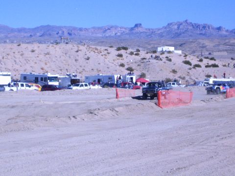 1990 Ford Bronco - dark off-road vehicle runs the desert course past RV-filled pits and orange fencing, with rocky peaks behind.