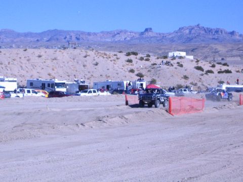 1990 Ford Bronco - boxy dark off-road vehicle runs the desert course beside orange fencing, with RVs, trucks and mountains behind.