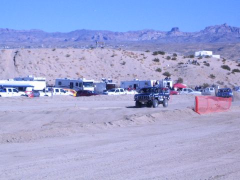 1990 Ford Bronco - dark off-road vehicle runs the desert course beside orange fencing, with RVs, support trucks and mountains behind.
