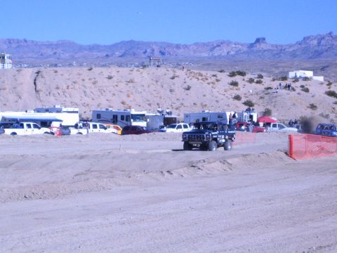 1990 Ford Bronco - dark off-road race truck rolls along a graded sand course lined with RVs and trucks, desert mountains in the distance.