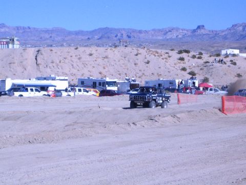 1990 Ford Bronco - blue off-road truck travels a packed-dirt desert course past a row of RVs and pickups under a clear blue sky.