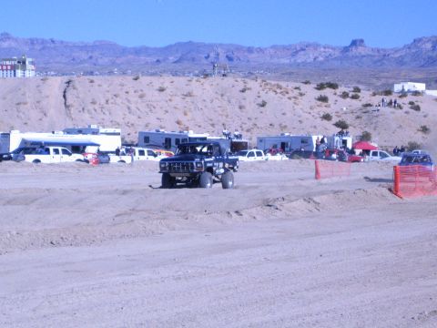 1990 Ford Bronco - dark lifted off-road truck drives toward the camera on a sandy desert race course flanked by RVs and orange fencing.