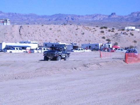 1990 Ford Bronco - dark off-road truck rounds a graded desert track, with parked RVs, pickups and a mountain ridge in the background.
