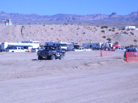 1990 Ford Bronco - dark lifted off-road truck travels a wide sandy desert race course past RVs and a red support vehicle.
