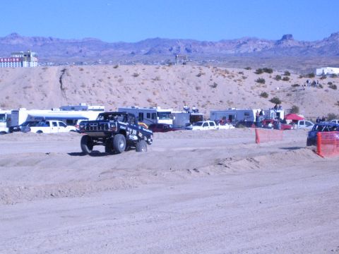 1990 Ford Bronco - dark off-road truck on oversized tires crosses a desert dirt track, with RVs and a casino on the hill behind.