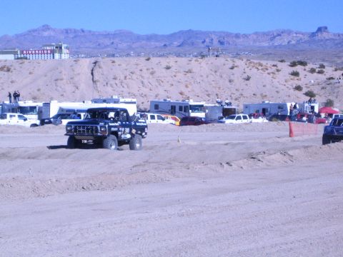1990 Ford Bronco - dark off-road race truck drives a desert dirt course, parked RVs and a hilltop casino visible in the background.