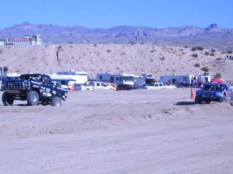 1990 Ford Bronco - dark number 300 off-road truck and a blue race truck run a sandy desert course lined with RVs.