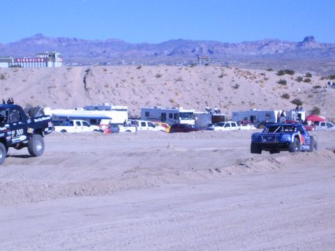 1990 Ford Bronco - number 300 dark off-road truck and a brightly liveried off-road truck run a desert course past parked RVs.