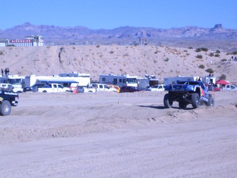 1990 Ford Bronco - dark race truck at left and a brightly liveried lifted off-road truck run a sandy desert race course with mountains behind.